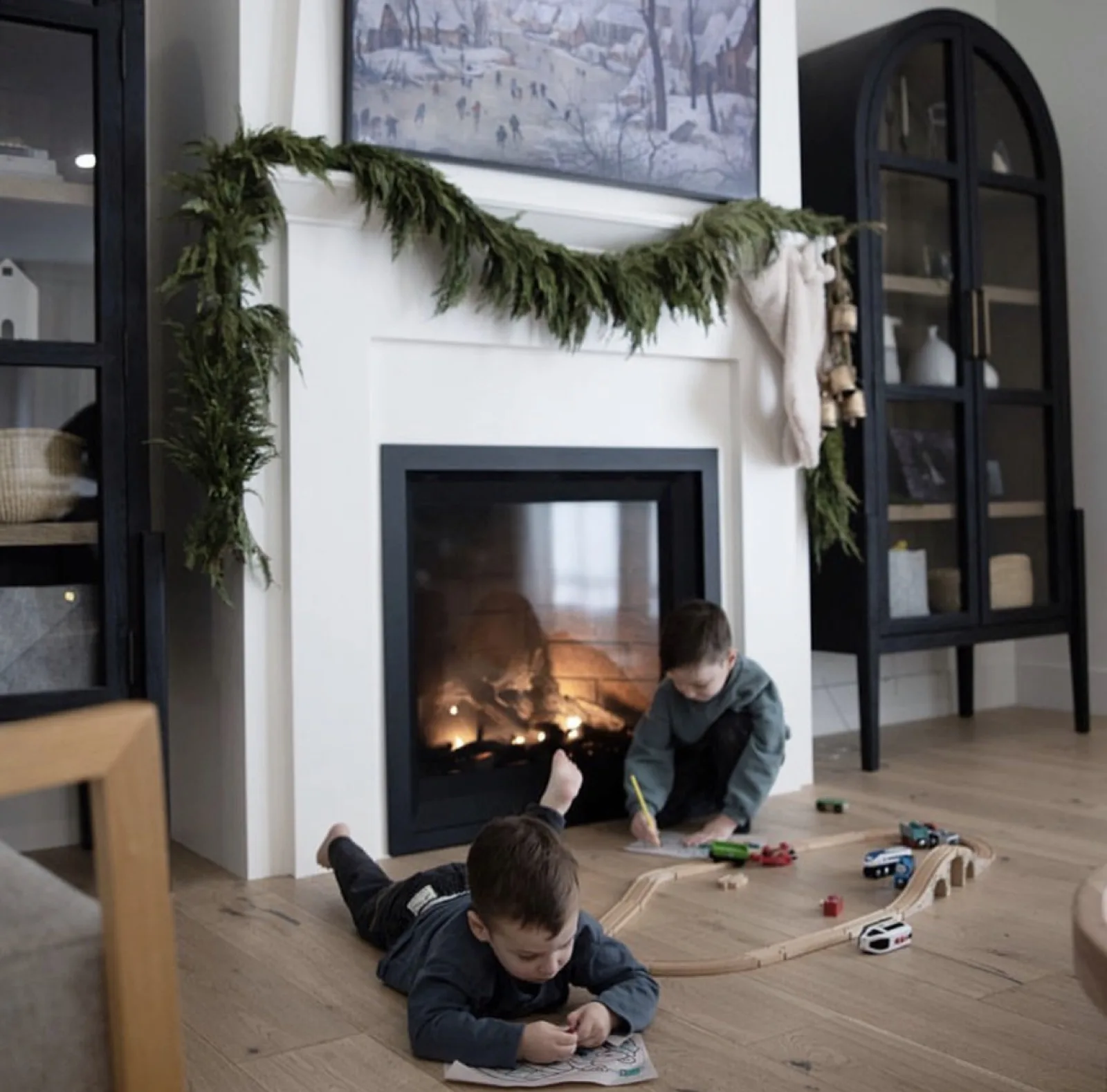 Two kids playing with a train set on the floor beside a burning see-through fireplace