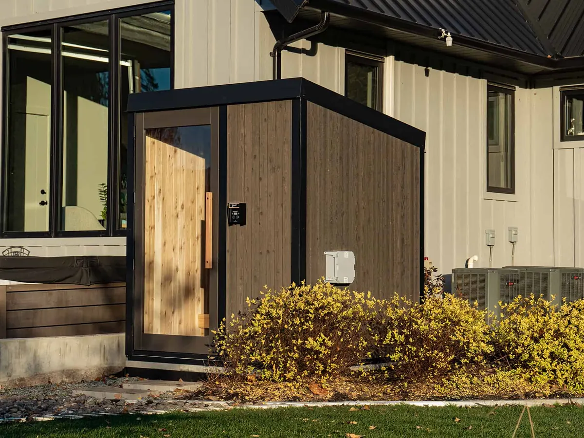 The Visscher Nature Series sauna, with Blacksmith cedar siding and a glass door, set beside a house